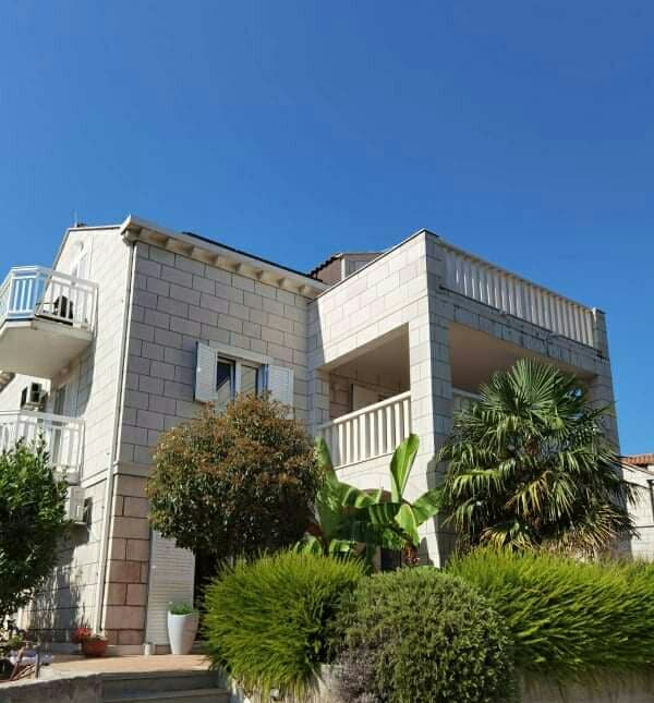 The exterior of the apartment building is crafted from light-colored stone, featuring a balcony on the upper level. Lush greenery, including palm trees and shrubs, surrounds the entrance, creating a serene environment. A clear blue sky serves as the backdrop, enhancing the inviting appearance of the structure.