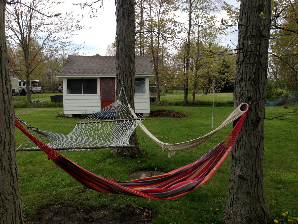 A vibrant hammock lies suspended between two trees, offering a space for relaxation. The grass below is well-maintained and green, while a rustic bunkie is visible in the background. Nearby trees and open space contribute to a serene outdoor environment.