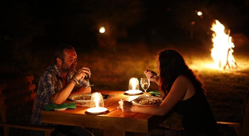 An outdoor dining setup is featured, with a wooden table illuminated by small lights. Two guests enjoy their meal, with plates and glasses visible. A cozy campfire burns nearby, providing warmth and a flickering ambiance in the natural surroundings.
