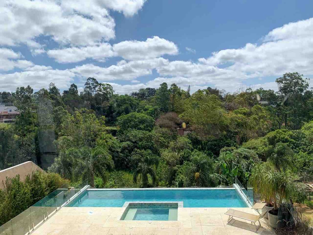 A serene view of lush greenery is observed from the edge of an infinity pool. The clear water of the pool reflects the bright sky, while surrounding palm trees add to the tranquil ambiance. Comfortable lounge chairs are positioned nearby for relaxation.
