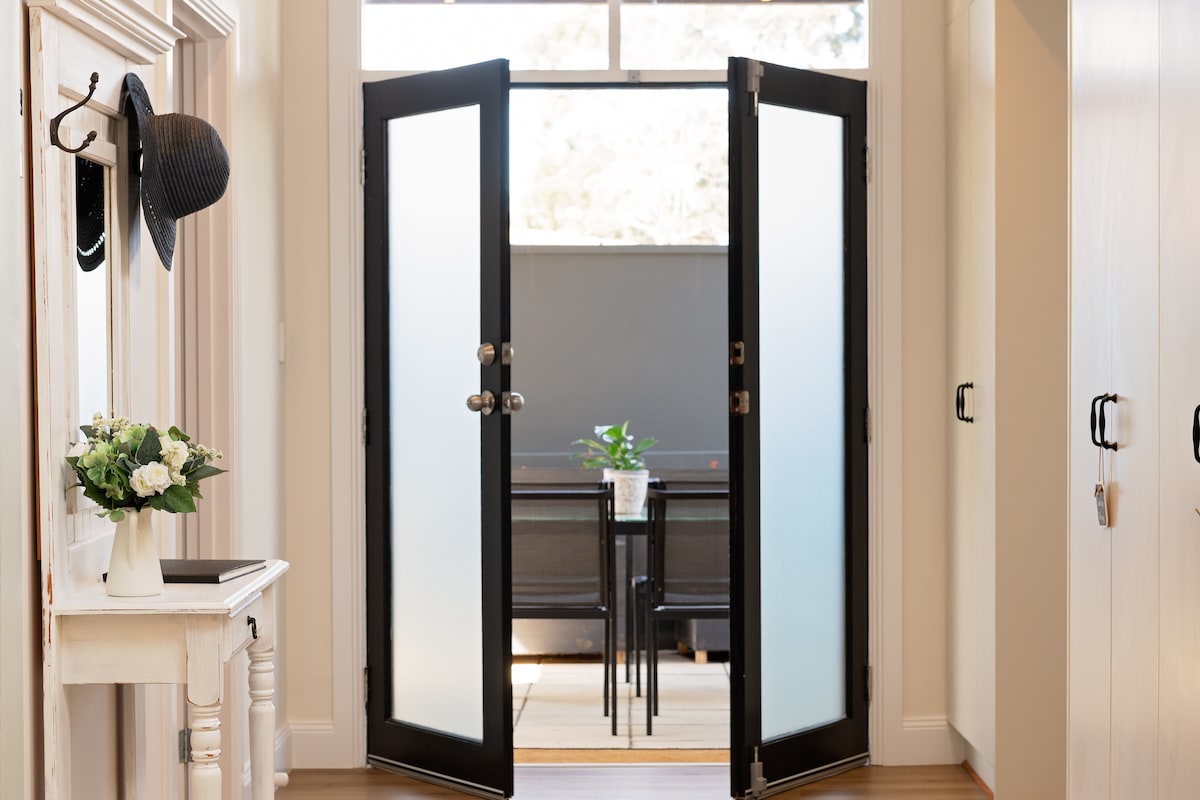 Double black doors, featuring frosted glass panels, open into a bright indoor space. A small dining table with chairs is visible in the background, alongside a potted plant. Sunlight filters through the large window, enhancing the warm atmosphere of the entrance.