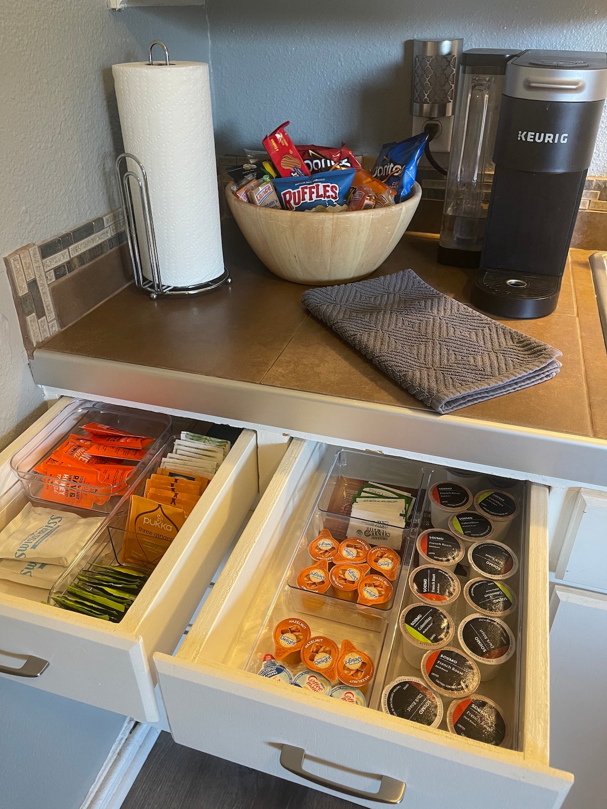 A kitchen area displays a countertop with a Keurig coffee maker, paper towels, and a bowl filled with snacks. Two drawers are partially open, revealing various coffee pod options and condiments. A textured gray towel is neatly placed next to the coffee maker.