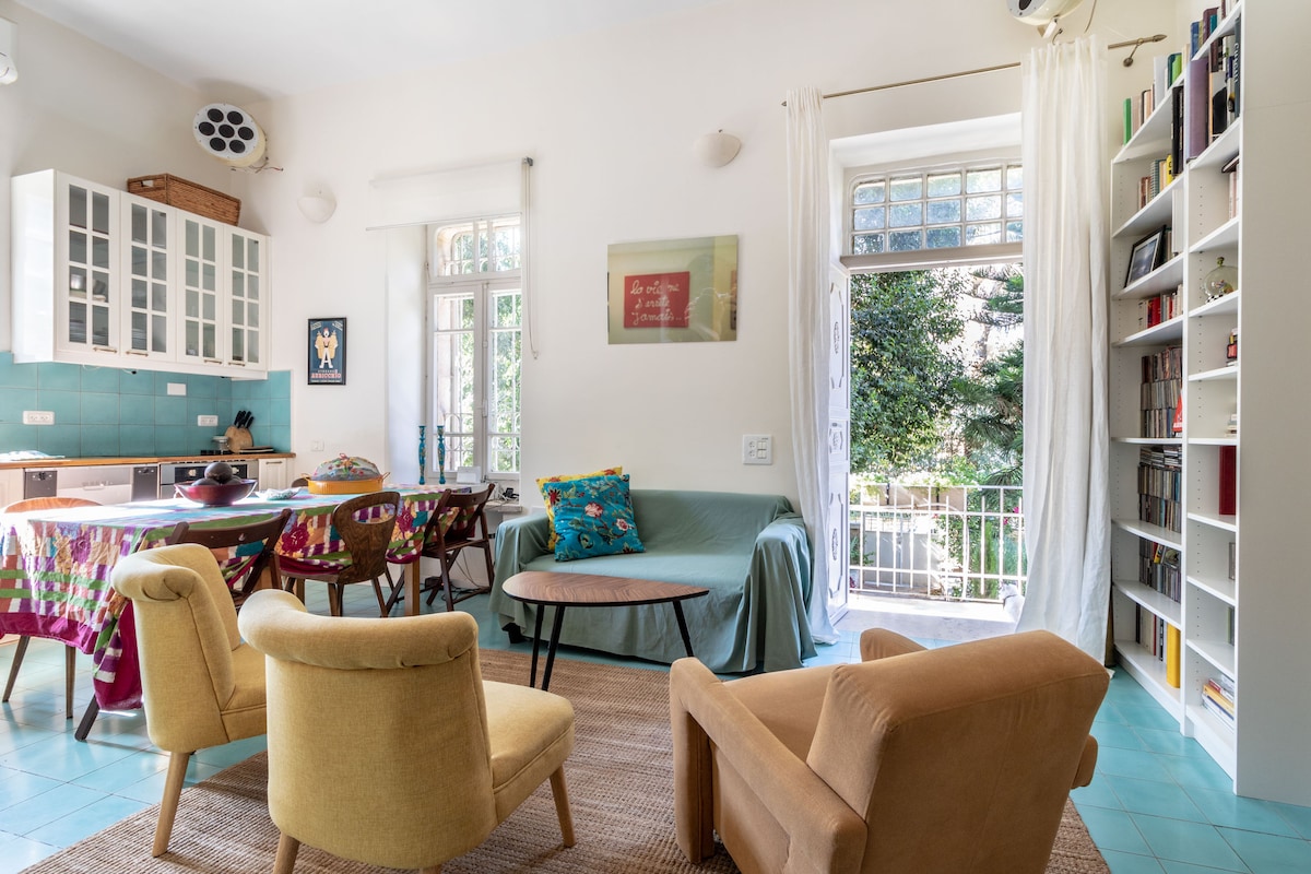 A bright living area is enriched by large windows that bring in natural light. Comfortable seating is arranged around a low table, while the kitchen features white cabinetry and colorful accents. A vibrant rug adds warmth to the space, which opens to a view of the garden.
