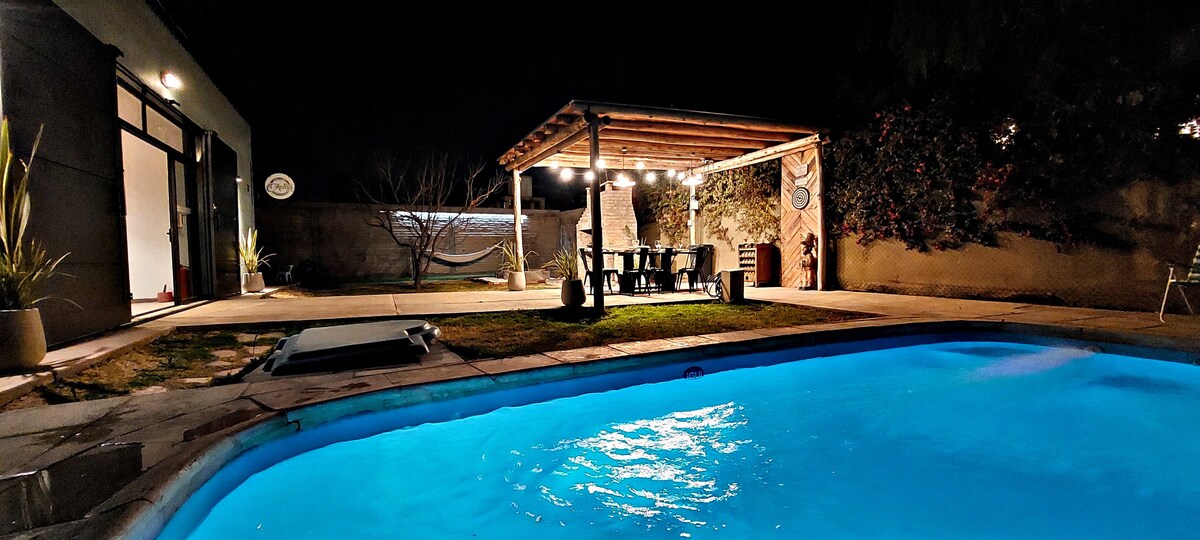 The outdoor area features a shimmering blue pool, complemented by a wooden pergola that houses a dining table and chairs. Soft lighting enhances the space, while surrounding greenery and a grassy patch create a serene ambiance for evening gatherings.