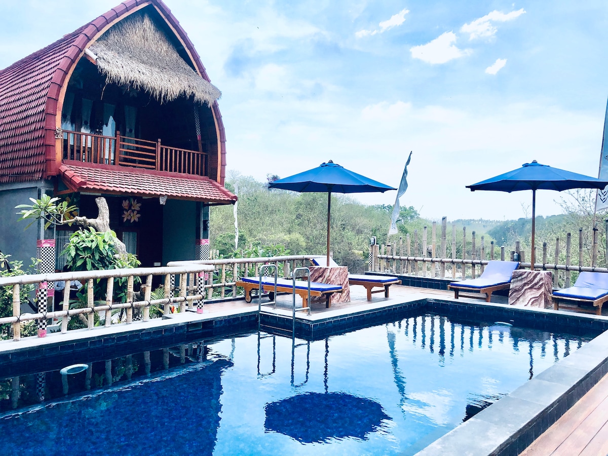A tranquil swimming pool is framed by wooden deck chairs and umbrellas, offering a scenic view of the surrounding landscape. The inviting pool reflects the blue sky above, while a traditional hut with a thatched roof and private balconies adds charm to the serene setting.
