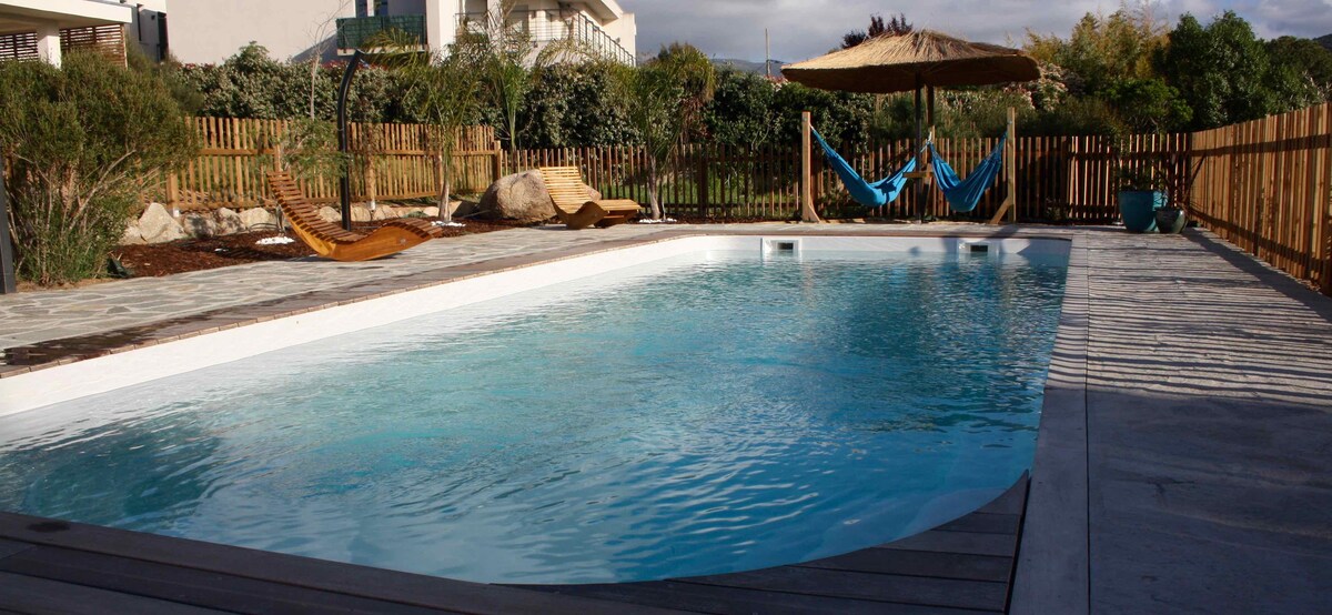 A clear swimming pool is surrounded by a spacious deck and wooden fencing. Sun loungers and a shaded area with a thatched roof add to the inviting outdoor space. Lush greenery can be seen in the background, enhancing the tranquil setting.