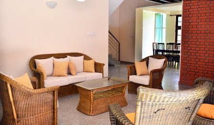 A comfortable seating area is highlighted by a rattan sofa and armchairs, arranged around a glass coffee table. Natural light filters in through large windows, illuminating the space with a relaxed and inviting ambiance.