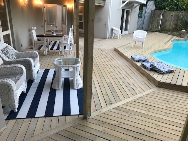 An outdoor deck area is seen featuring a rectangular pool, accompanied by white wicker seating and a small table. A dining set with four chairs is positioned nearby on a striped rug. Two lounge chairs are arranged on the deck, fostering a relaxed environment.