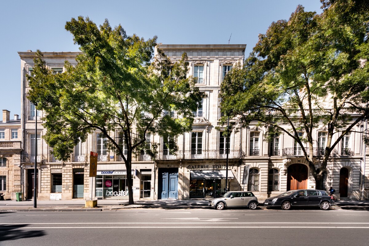 The exterior of a historic stone building from the 18th century is visible, showcasing elegant architecture with large windows and a classic façade. Lush trees line the street, providing shade, while parked cars are arranged along the curb.