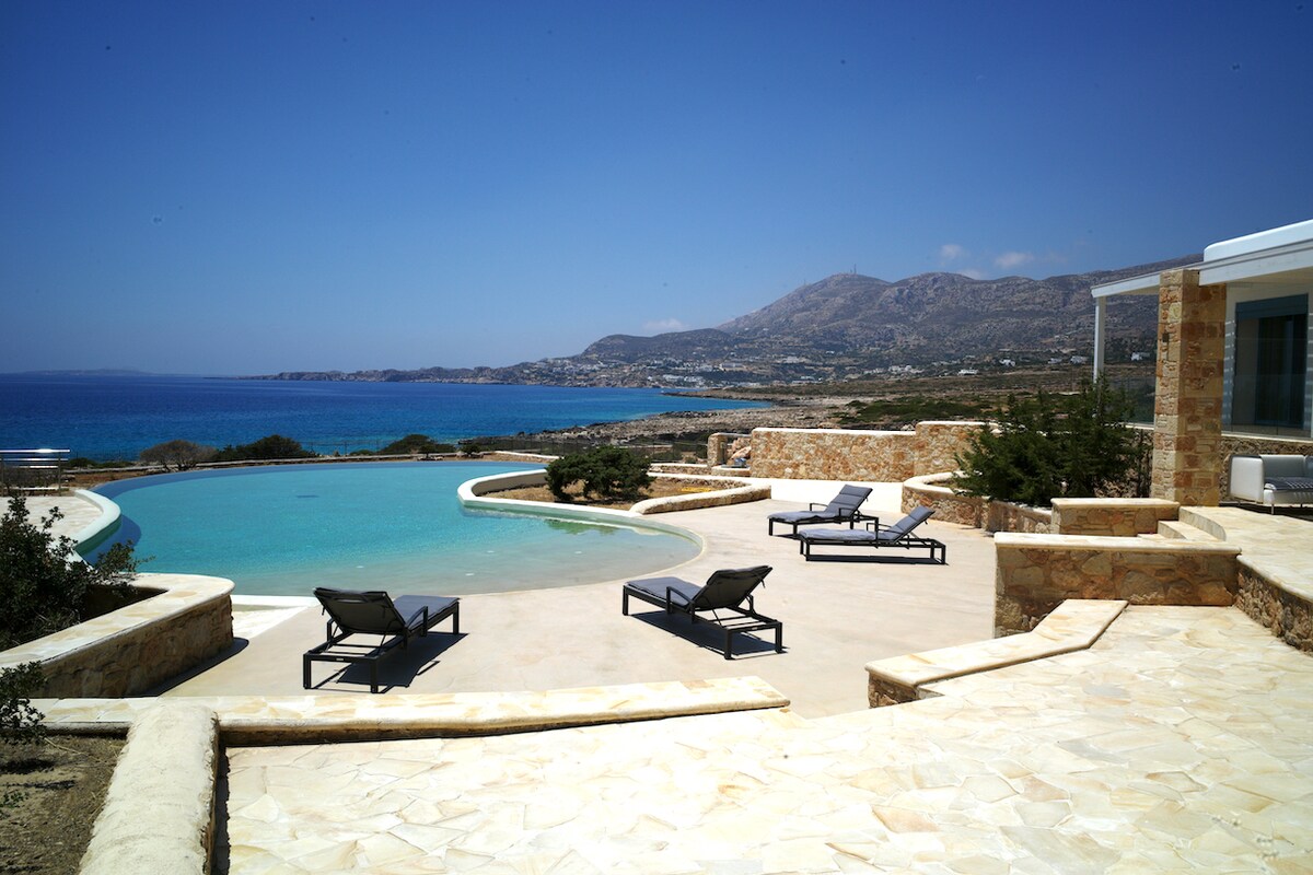 A spacious outdoor area features a curved swimming pool surrounded by sun loungers. The patio is adorned with natural stone, leading toward a tranquil sea view. Rolling hills and a blue sky create a serene backdrop for the setting.