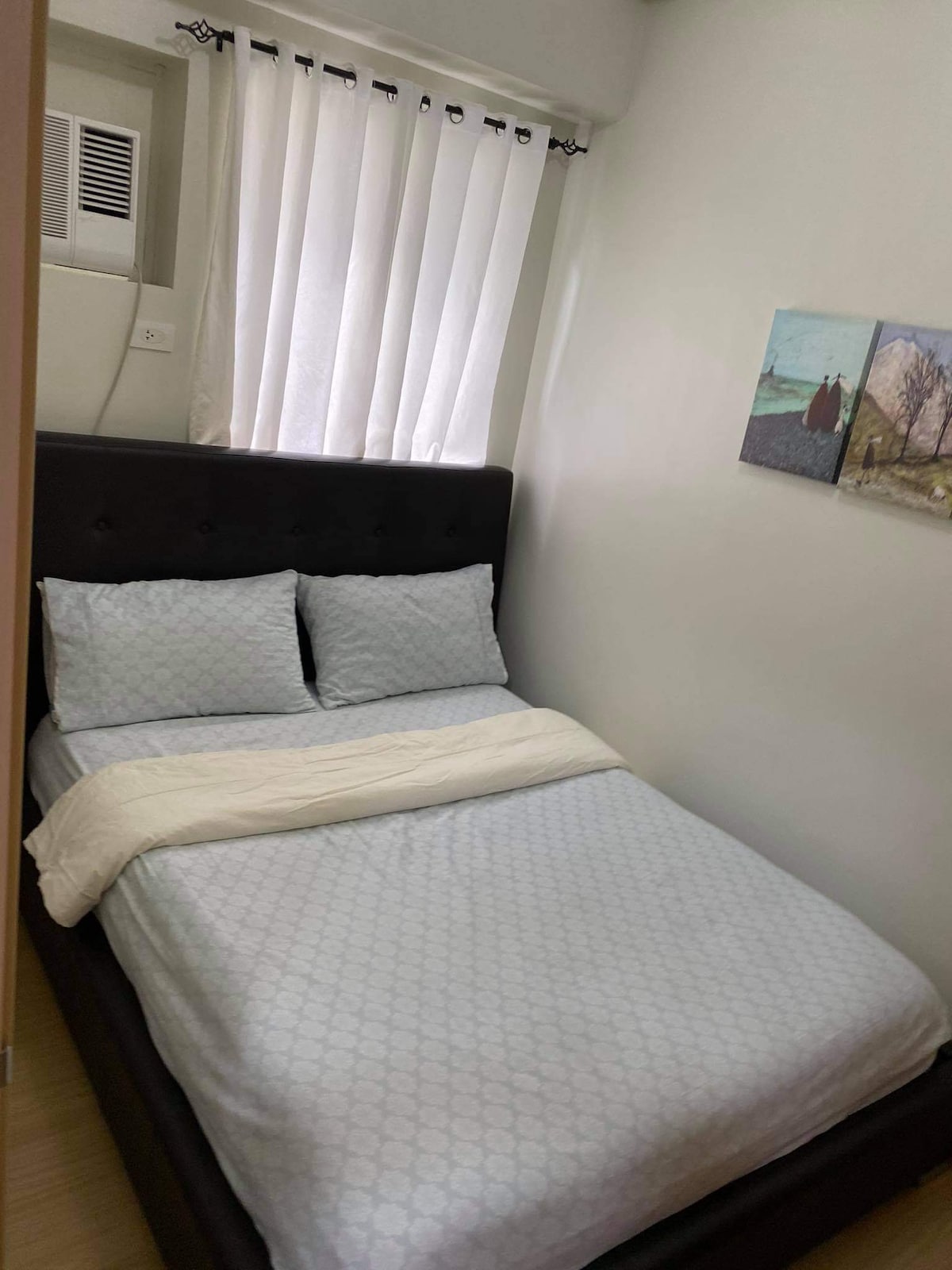A compact bedroom is presented with a dark upholstered bed featuring light blue bedding and two pillows. Natural light enters through vertical sheer curtains. Two framed artworks are displayed on the wall adjacent to the bed, adding a touch of color to the minimalist decor.