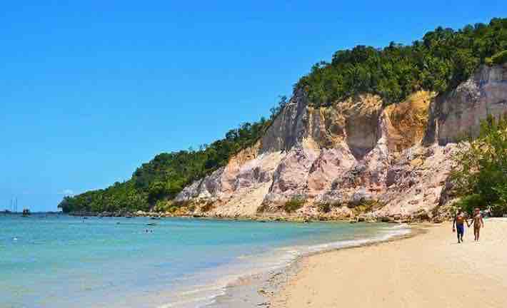 A sandy beach stretches along the shore, framed by towering cliffs adorned with greenery. Clear blue waters gently lap against the shoreline, while individuals can be seen walking along the beach, enjoying the serene landscape and the calming presence of nature.