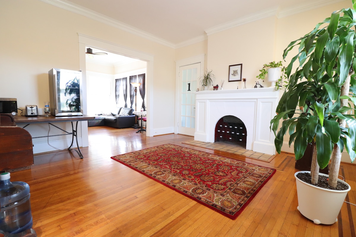 A spacious living area is accentuated by warm wooden floors and a decorative area rug. A prominent fireplace is flanked by houseplants, and a flat-screen TV is positioned on a table. Natural light streams in from large windows, creating a bright atmosphere.