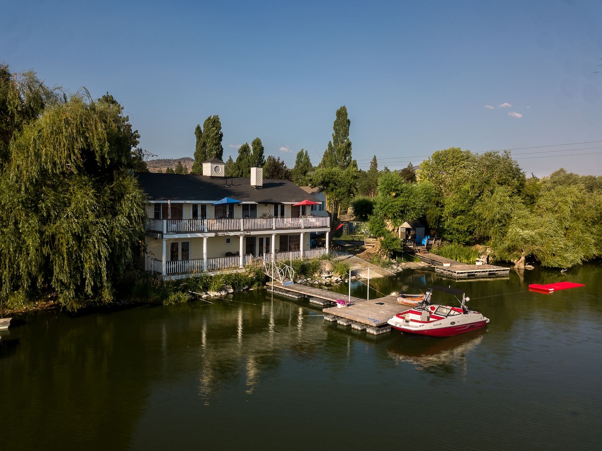 A spacious two-story home is positioned along the lakefront, featuring multiple decks ideal for enjoying the scenery. A dock with a boat is visible, and the surrounding lush greenery adds a natural touch. Calm waters reflect the clear sky, enhancing the serene atmosphere.