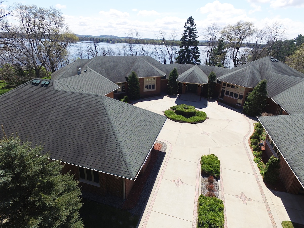 An aerial view captures the expansive layout of the mansion, displaying a circular driveway surrounded by landscaped greenery. The serene waters of Tainter Lake are visible in the background, complementing the peaceful environment of this lakeside retreat.