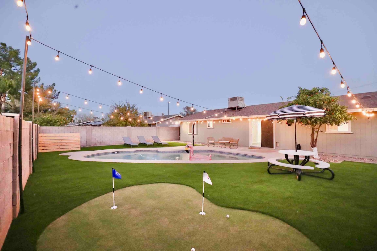 The backyard features a well-maintained lawn with a putting green, a sparkling swimming pool, and a picnic table. String lights are hung above, creating a warm ambiance for evening gatherings. Lounge chairs are positioned nearby, offering a relaxing spot to enjoy the outdoor space.