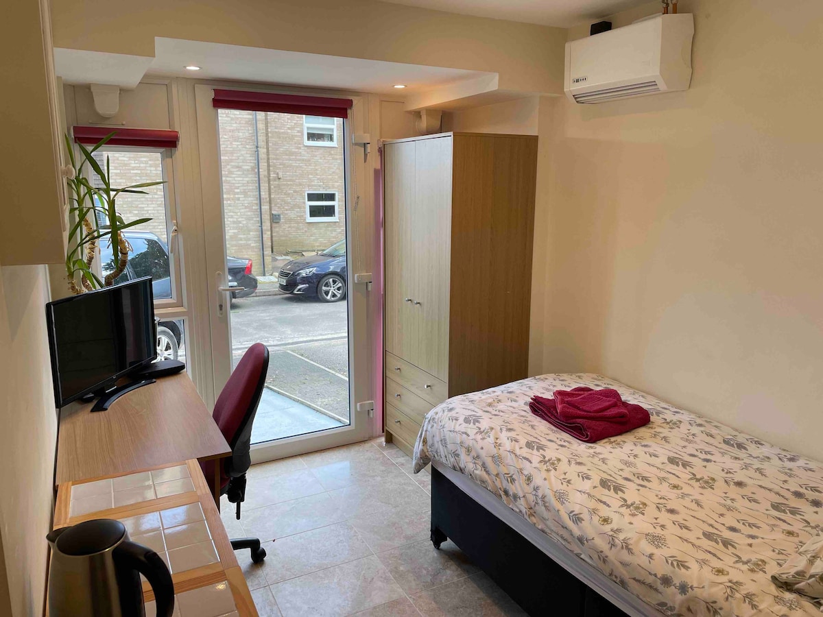 A neatly arranged room features a single bed with floral bedding, complemented by a red throw. A wooden desk with a chair is positioned near a window that provides natural light. A television is on the desk, alongside a kettle. Natural greenery is present in a nearby planter.