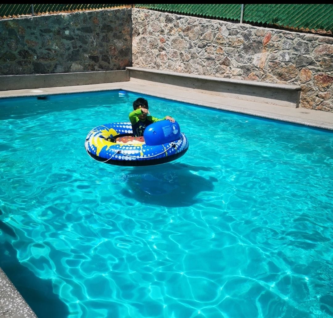 A child enjoys a colorful inflatable float in the center of a clear blue swimming pool. Surrounding stone walls provide privacy, while the water reflects sunlight, creating a bright and inviting atmosphere.