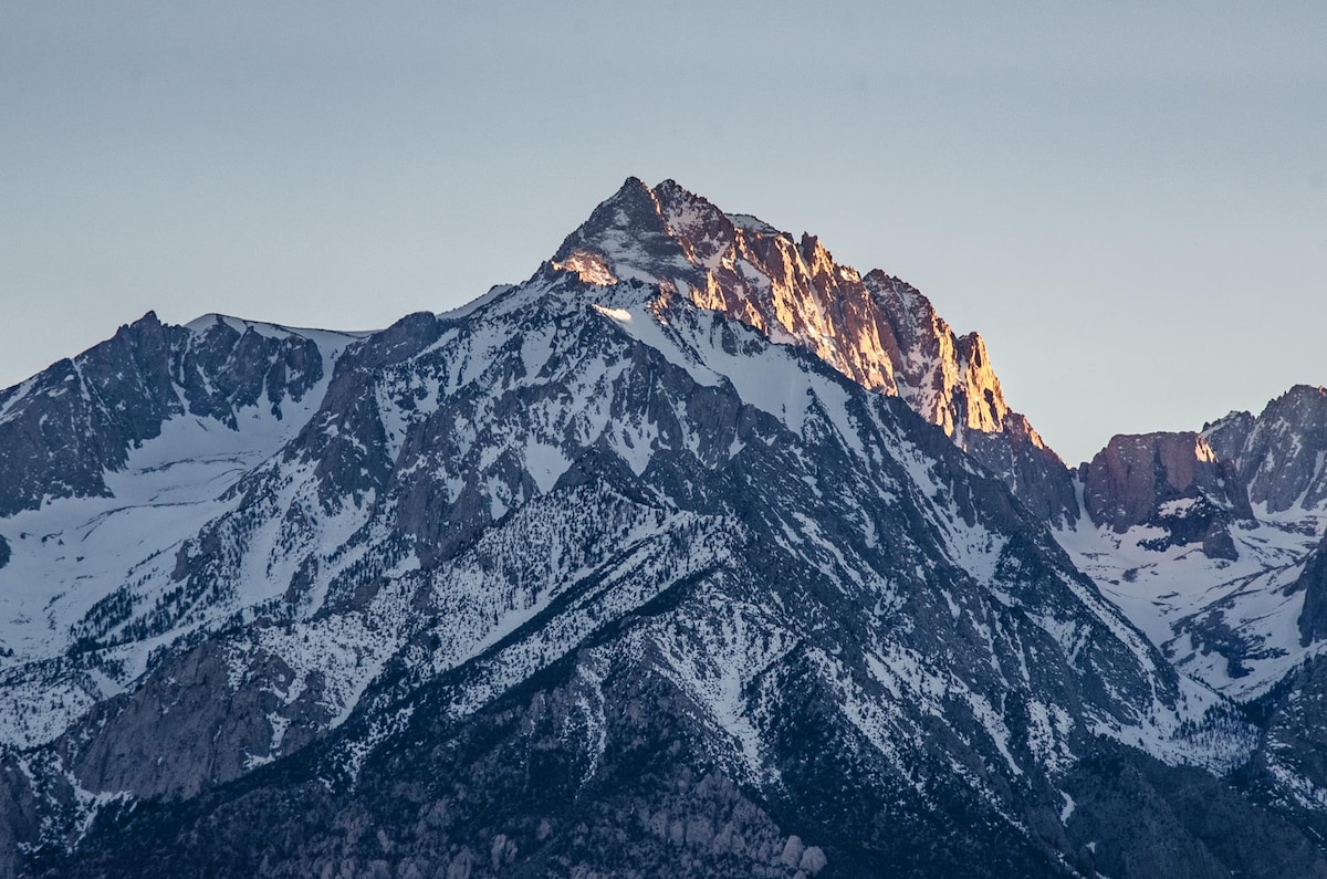A majestic mountain peak is captured in soft morning light, highlighting its snow-capped summit and rugged terrain. The surrounding landscape features a blend of rocky slopes and patches of green, creating a serene and natural environment.