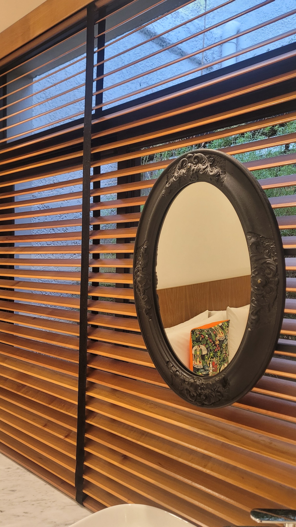 A modern wooden slat screen separates the sleeping area from the outside, allowing light to filter through. An oval, intricately designed mirror hangs on the wall, reflecting the warm tones of the wood. A subtle view of greenery can be seen through the slats.