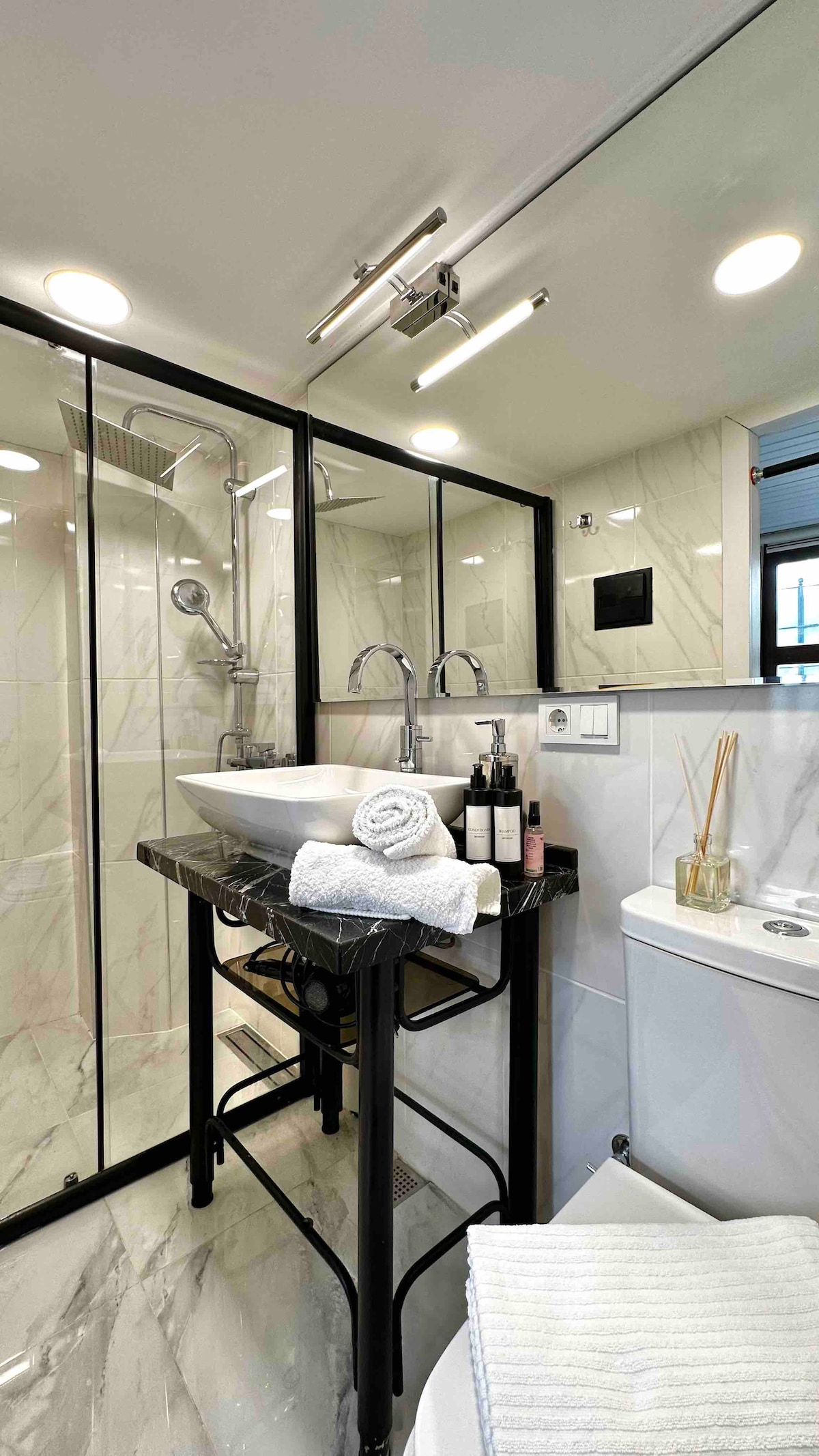 A modern bathroom is equipped with a glass shower enclosure and sleek marble accents. A stylish black sink with a marble top is placed atop a metal frame. Bottles and toiletries are neatly arranged alongside fresh towels, contributing to a clean and organized space.