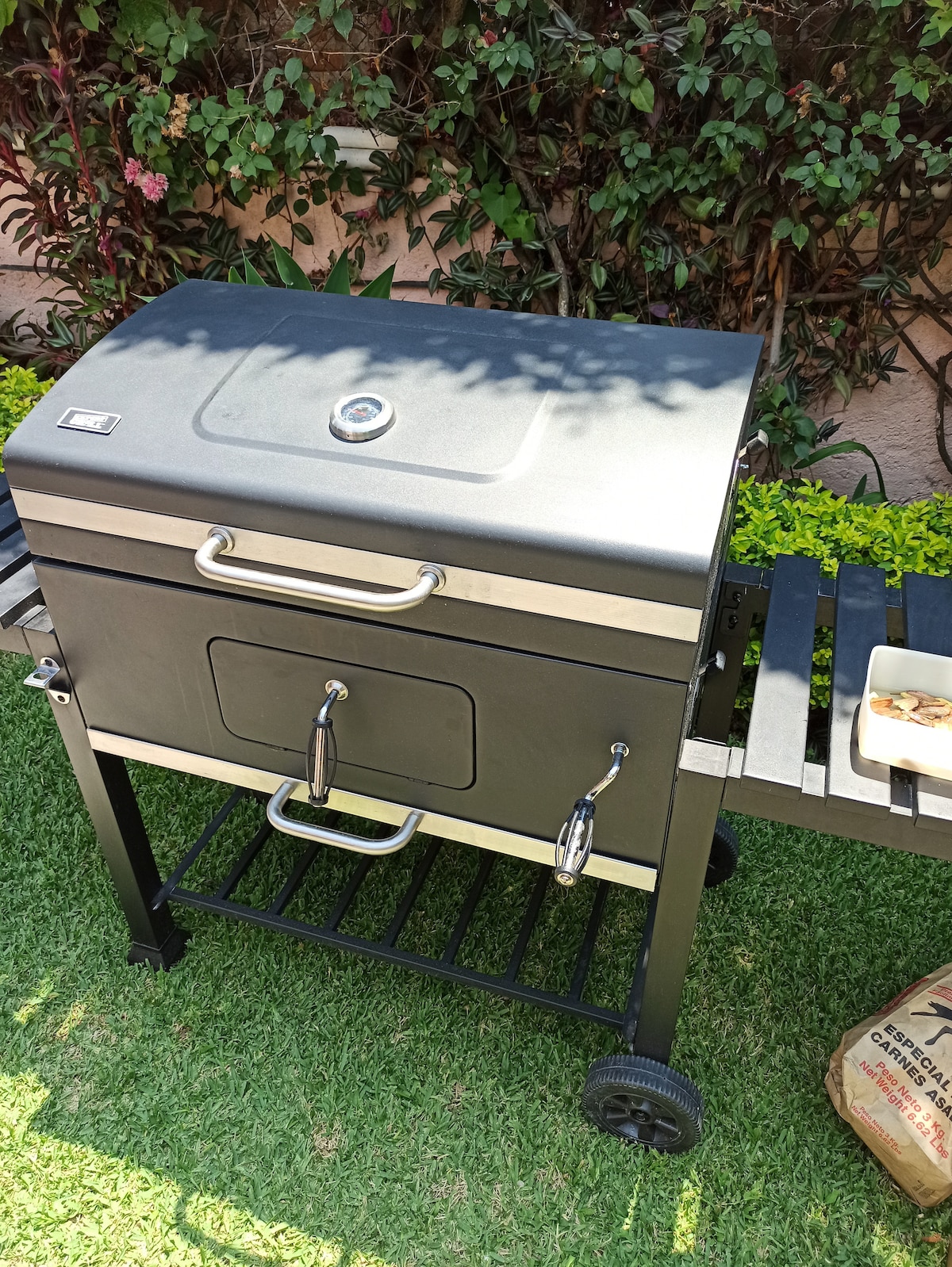 A charcoal grill is positioned on a black metal cart with wheels, situated on a patch of green grass. The grill features a smooth, grey top with a sturdy handle and two front-facing doors. Nearby, a wooden bench provides additional space for food preparation.