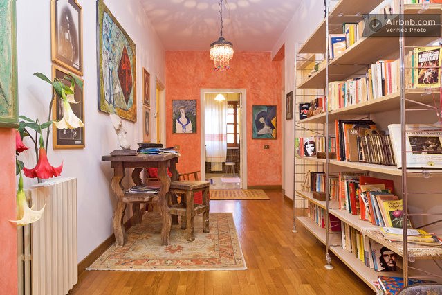 A hallway is illuminated by a chandelier, featuring a wooden table surrounded by decorative art pieces. A bookshelf filled with various books lines one wall, while warm tones and a patterned rug create a welcoming atmosphere.
