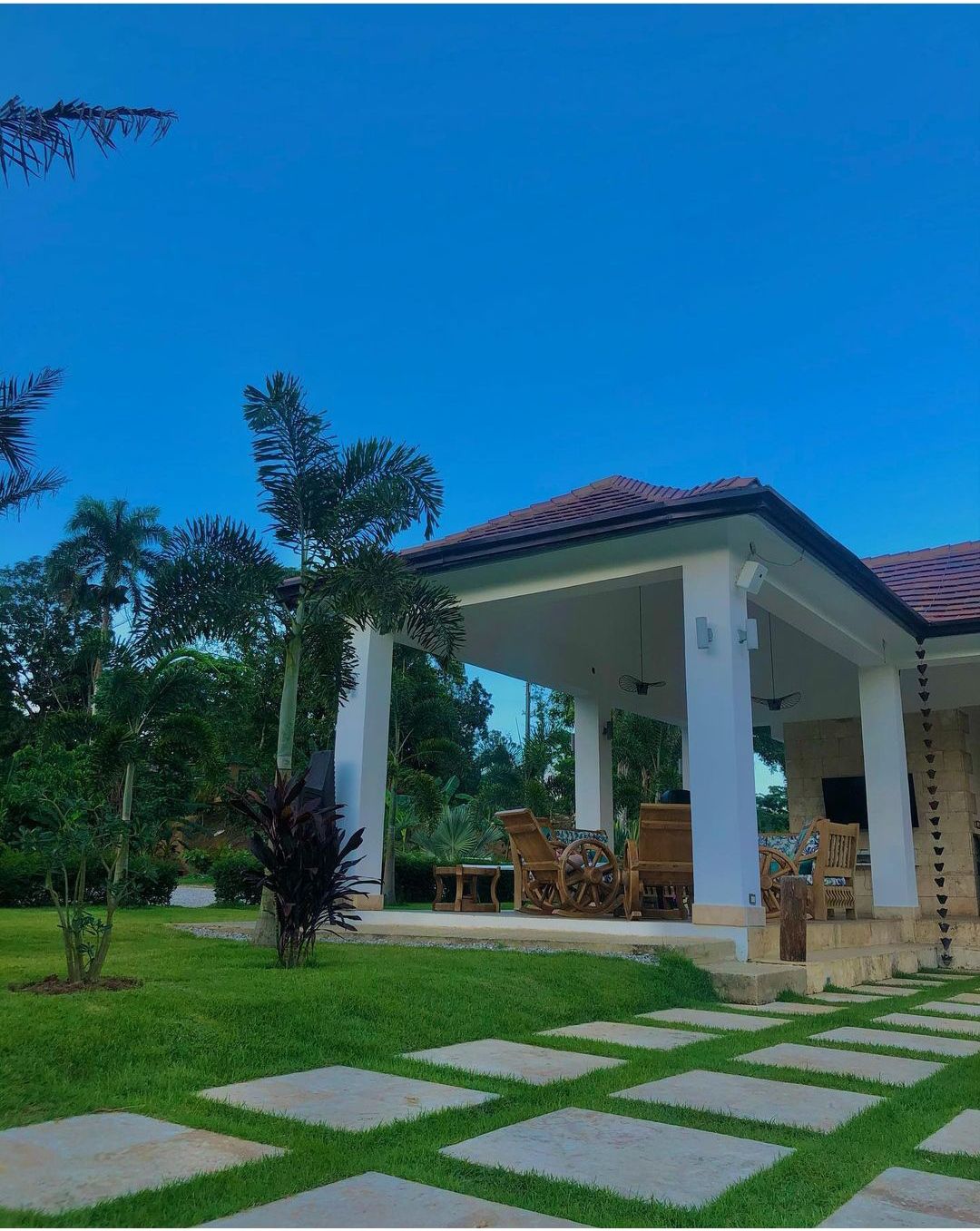 A covered patio is framed by tall white columns, featuring a wooden dining set with chairs. The surrounding area displays a well-maintained lawn with stepping stones leading towards lush greenery and palm trees, all under a clear blue sky.