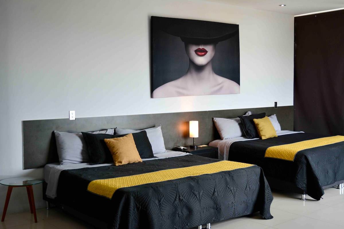 The room features two spacious beds dressed in neutral tones, complemented by decorative pillows. A modern nightstand with a lamp is situated between the beds. A large wall art piece adds character, and natural light enters from a nearby window covered by dark curtains.