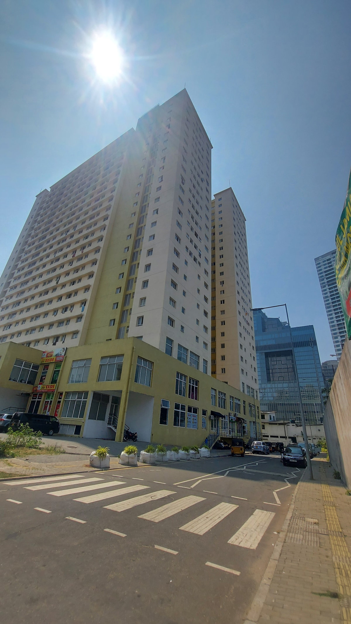 A tall apartment building is viewed from street level, showcasing a modern façade with a mix of light and dark shades. The surrounding area includes a pedestrian crosswalk and parked vehicles, with a bright sun illuminating the scene above.