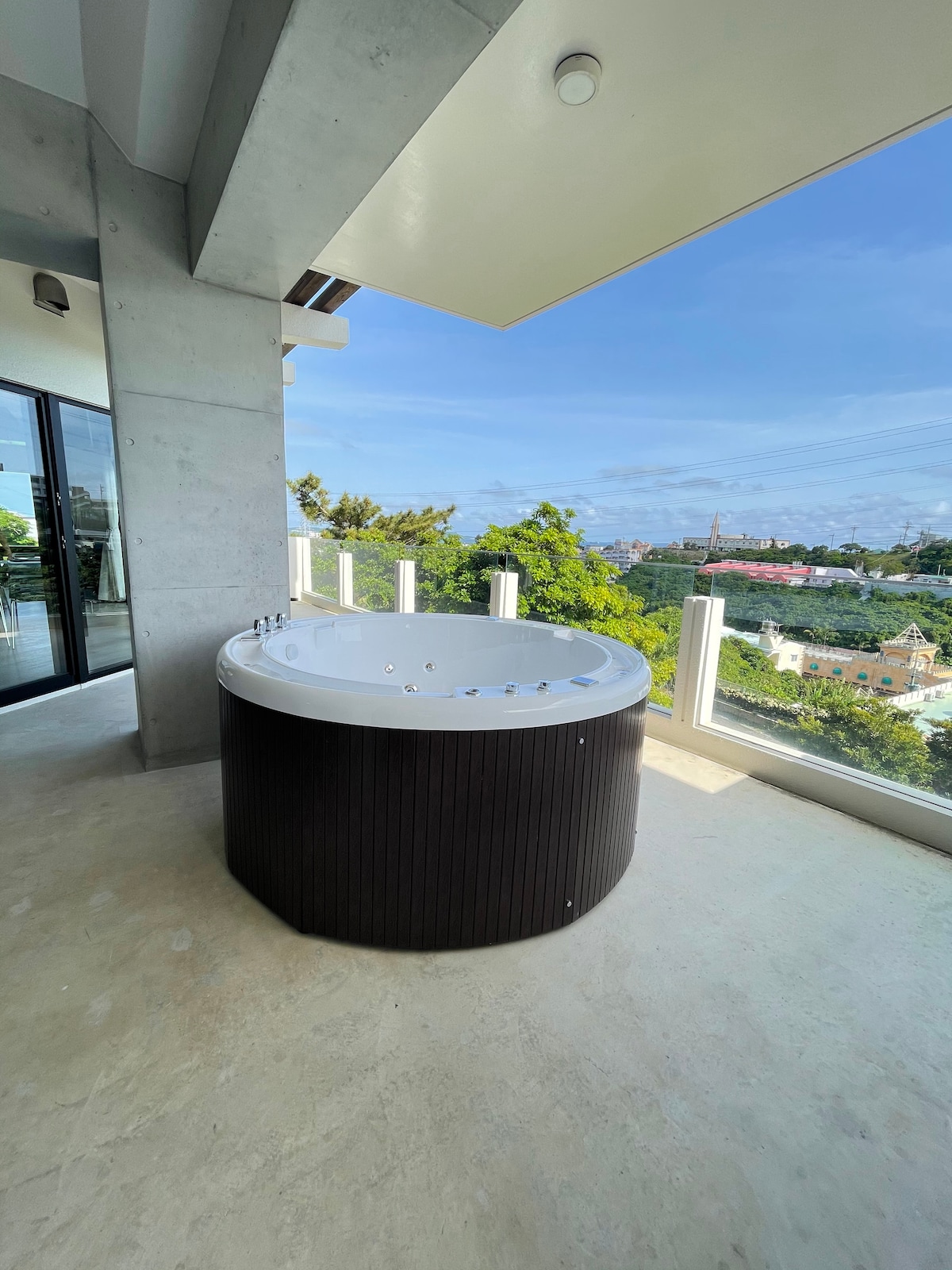 An outdoor terrace features a circular hot tub with temperature control settings, surrounded by a railing that offers views of the landscape. Bright blue skies are visible beyond, enhancing the inviting atmosphere of the space.