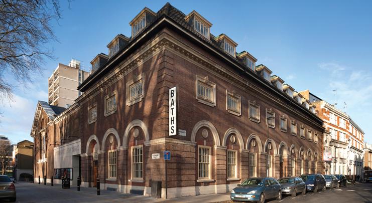 The exterior of Ironmonger Row Baths is showcased, featuring a classic brick facade with large arched windows. A prominent sign reading 'BATHS' is displayed on the building, while neatly parked cars line the street in front.