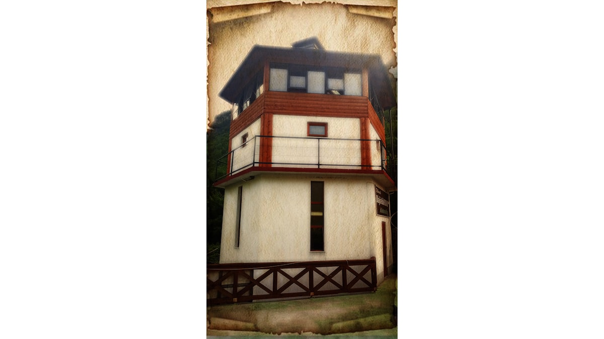 A multi-story building is presented, featuring a mix of wooden and light-colored wall elements. Large windows allow natural light to illuminate the interior, while a balcony extends from the upper level, offering views of the surrounding area. A wooden fence encloses the lower level.