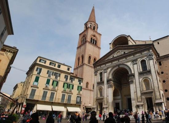 A historical square features a tall church tower with a pointed spire rising above nearby buildings. The stone facade of a grand church entrance is visible, surrounded by bustling pedestrians. Cafés and shops line the street, contributing to the vibrant atmosphere of the area.