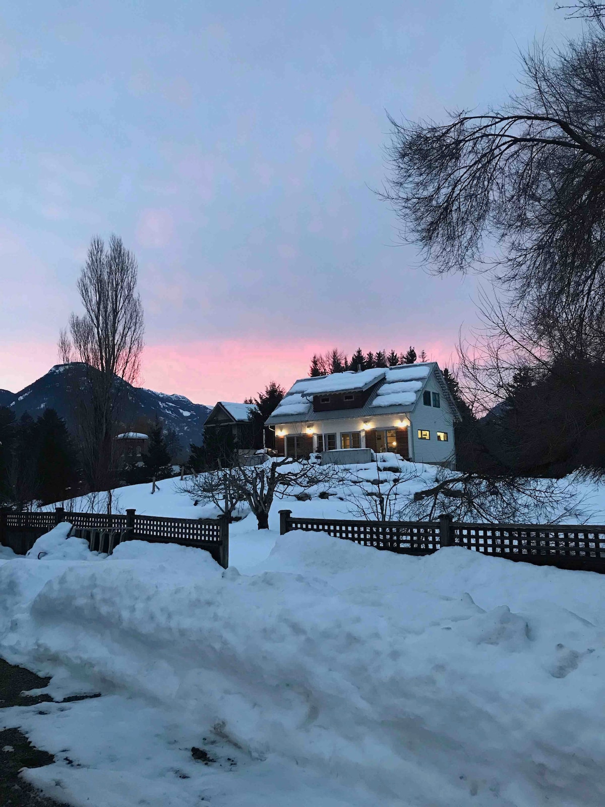 A modern chalet farmhouse is seen from a distance, surrounded by snow-covered ground. The soft glow of lights emanates from the windows, while a pink and purple sky reflects the sunset behind the mountainous backdrop.