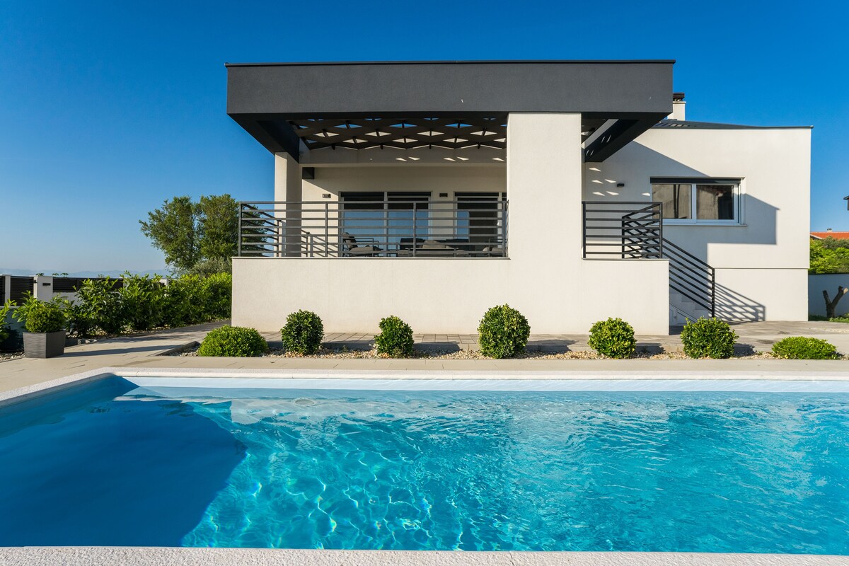 The exterior of a modern villa is captured, showcasing a spacious terrace with railing and lounge furniture. A clear swimming pool occupies the foreground, reflecting blue skies. Lush greenery is observed around the property, providing a serene backdrop.