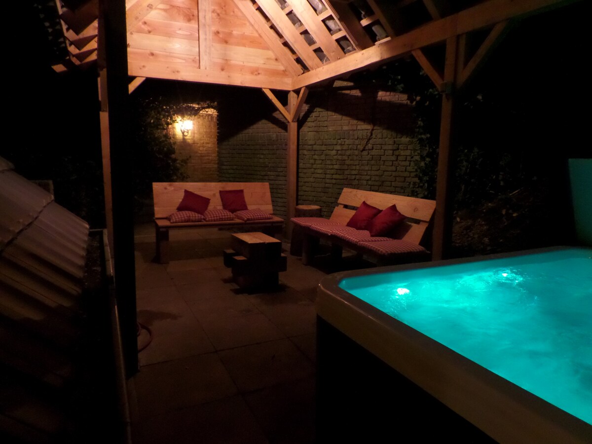 A private outdoor area is featured, showcasing a hot tub with glowing blue water. Wooden benches are arranged with cushions, providing a cozy seating space. Soft lighting from a wall lamp enhances the ambiance, while the surrounding greenery creates a secluded atmosphere.