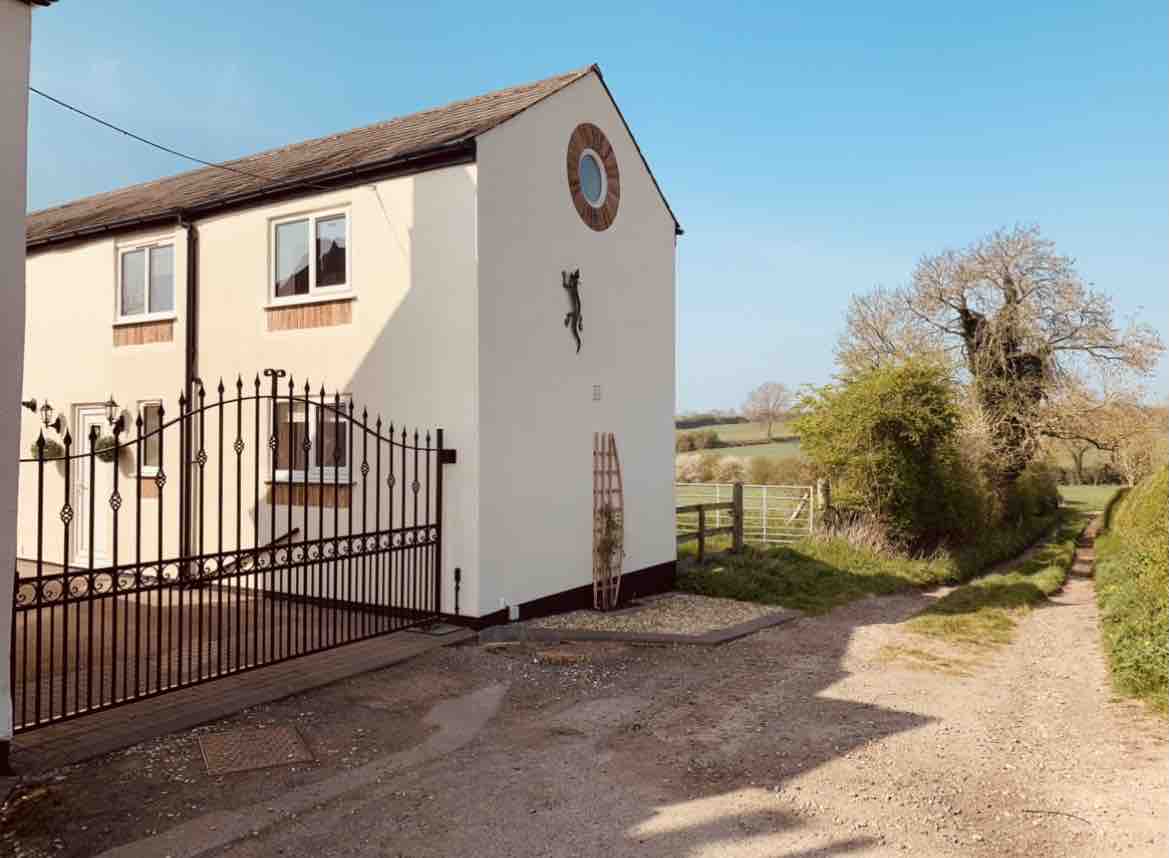 The image shows a two-story building set beside a gravel path leading through a countryside gate. The exterior features a light-colored facade and large windows, with greenery visible in the surrounding area, offering a serene rural atmosphere.