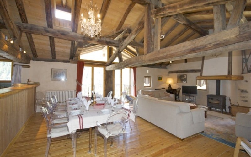 An open-plan living and dining area features rustic wooden beams and a chandelier overhead. A large dining table is set with tableware, while comfortable seating is arranged around a cozy wood-burning stove. Light pours in through large windows, creating a warm and inviting space.