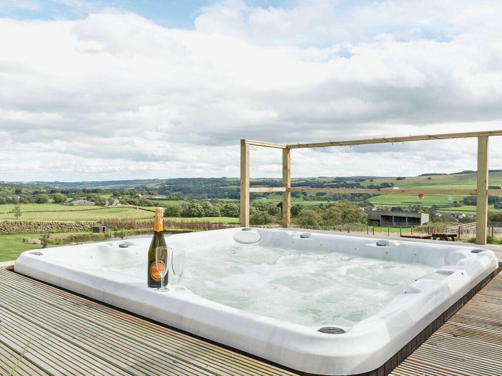 A private hot tub is positioned on a wooden deck, offering scenic views of the surrounding landscape. A bottle of champagne and a glass are placed on the side, highlighting a relaxing atmosphere. The sky features a mix of clouds and blue, enhancing the outdoor experience.