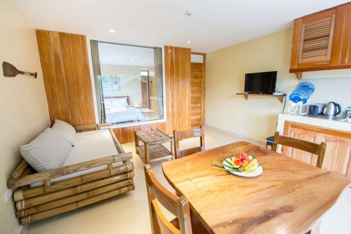 A cozy living area features a bamboo sofa and a wooden dining table surrounded by chairs. A kitchenette is visible in the background, equipped with essential appliances. Natural light fills the space through a large window, enhancing the warm wooden accents.