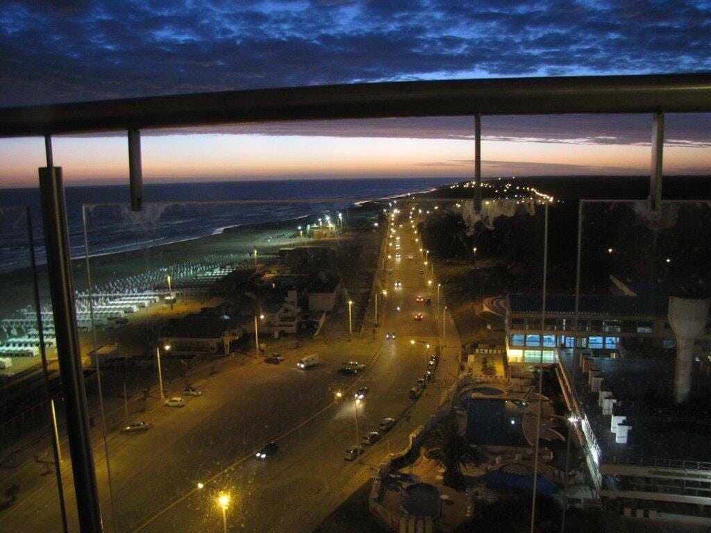 A sweeping view of the coastline is captured from a balcony, showing the ocean's horizon under a twilight sky. Streetlights illuminate the road leading to the beach, while the serene waves can be seen in the distance.