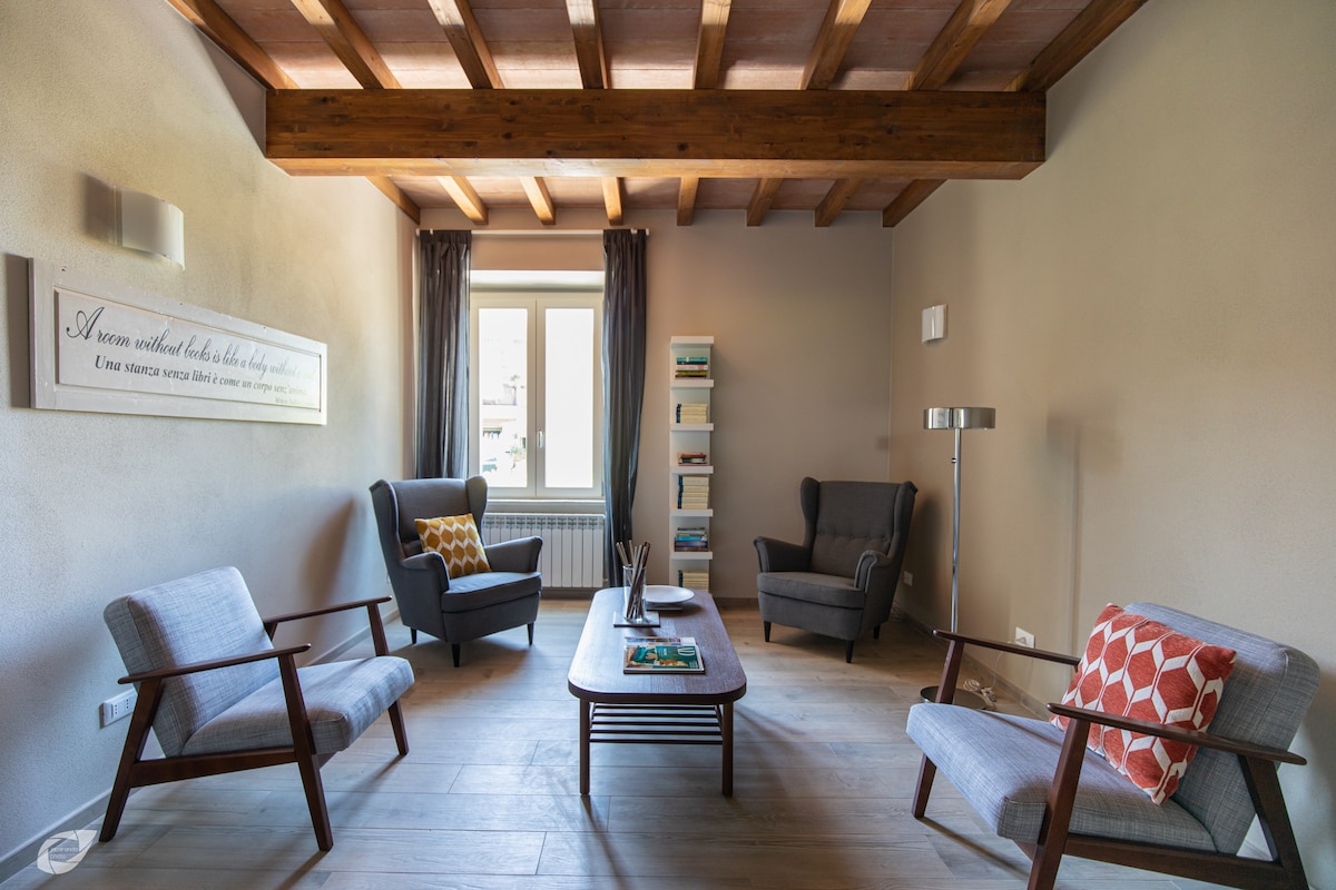 A cozy reading area is presented, featuring two armchairs and two upholstered chairs arranged around a wooden coffee table. A bookshelf displays various books, while natural light filters in through the window, enhancing the serene environment. Wooden beams add character to the ceiling.