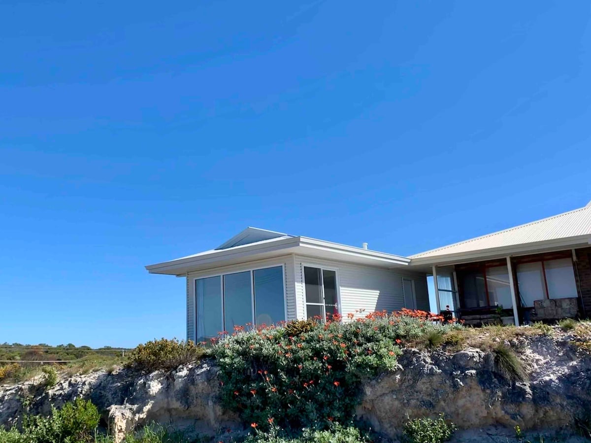 A single-story structure with large glass windows is set against a clear blue sky. Colorful flowers and green plants are visible in the foreground, enhancing the natural landscape. The building features a sloped roof and is surrounded by rocky terrain, showcasing the coastal environment.