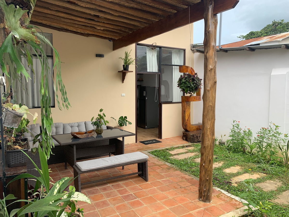 An intimate outdoor seating area features a shaded table with comfortable seating, surrounded by lush greenery. A well-kept garden path leads to an entrance with modern appliances visible inside the home. Natural light filters through large windows, creating a welcoming atmosphere.