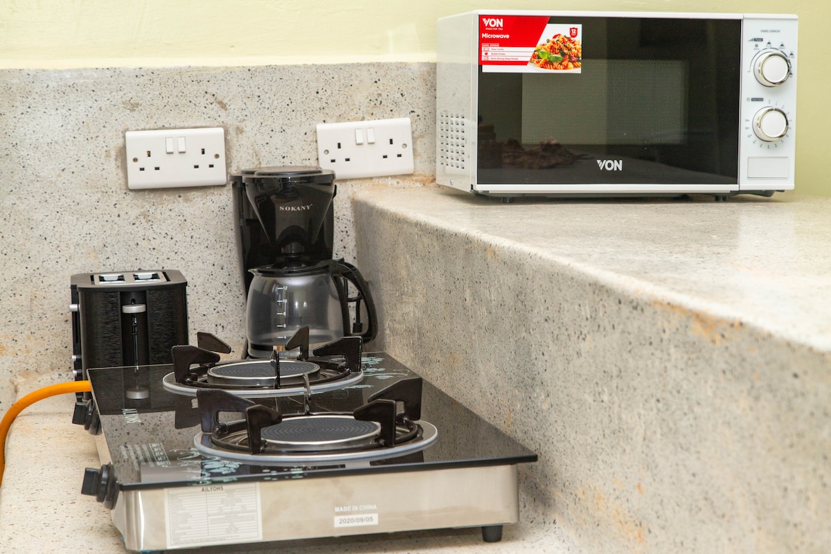 A modern kitchen workspace is shown, featuring a two-burner gas cooktop beside a microwave. A coffee maker is positioned near the cooktop, with electrical outlets visible above the countertop. The surface is made of a light-colored stone, offering a clean and functional cooking area.