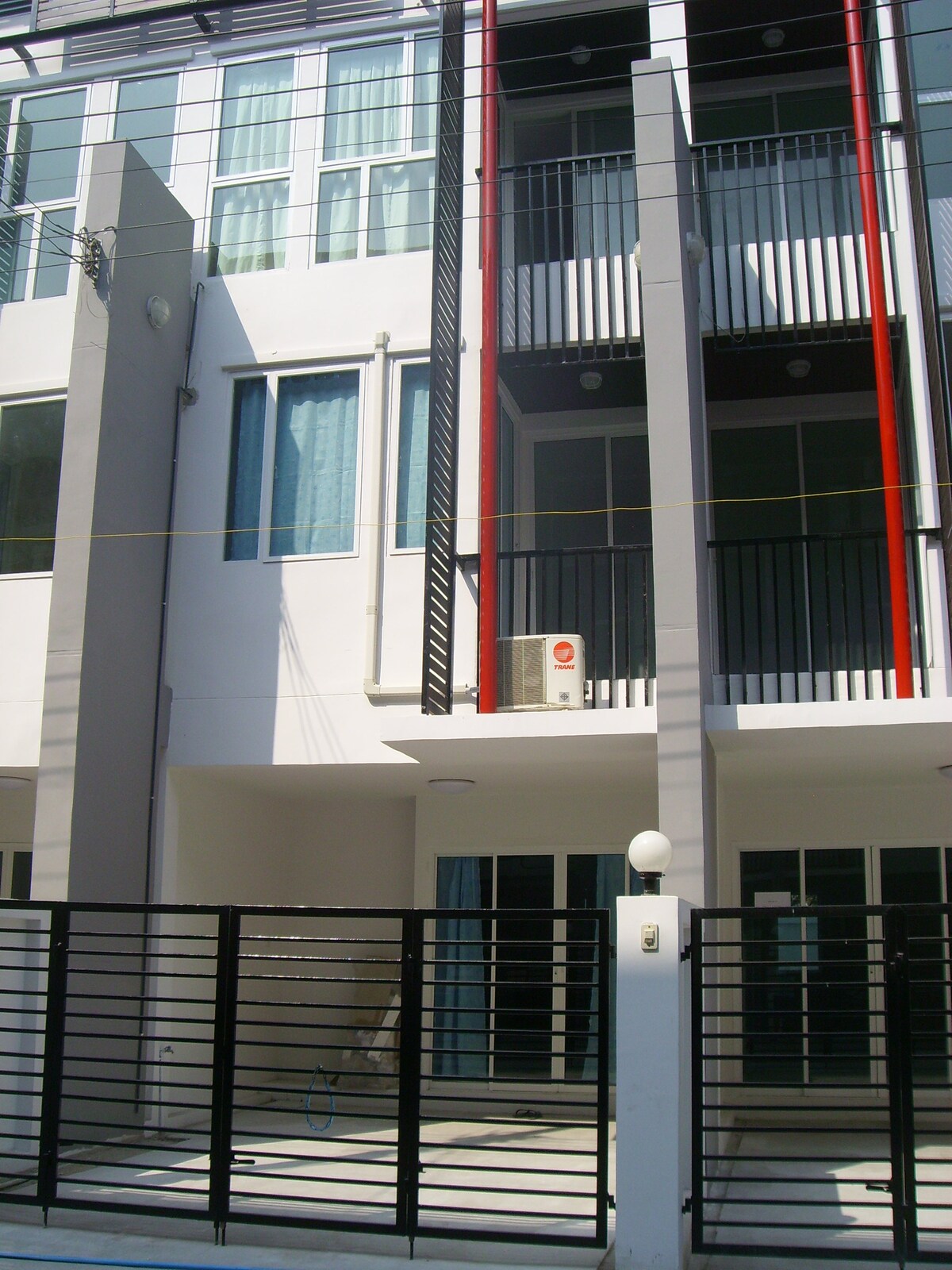 A modern multi-story building showcases a clean façade with sleek lines. Large windows with light blue curtains allow natural light to fill the interior. An air conditioning unit is mounted on the side, and a gated entrance adds privacy and security.