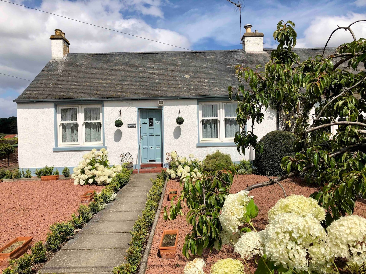 The cottage exterior features a white facade with blue accents, framed by well-maintained gardens. A pathway leads to the bright blue front door, flanked by symmetrical windows and a welcoming entry. Floral arrangements add a touch of greenery to the vibrant scene.