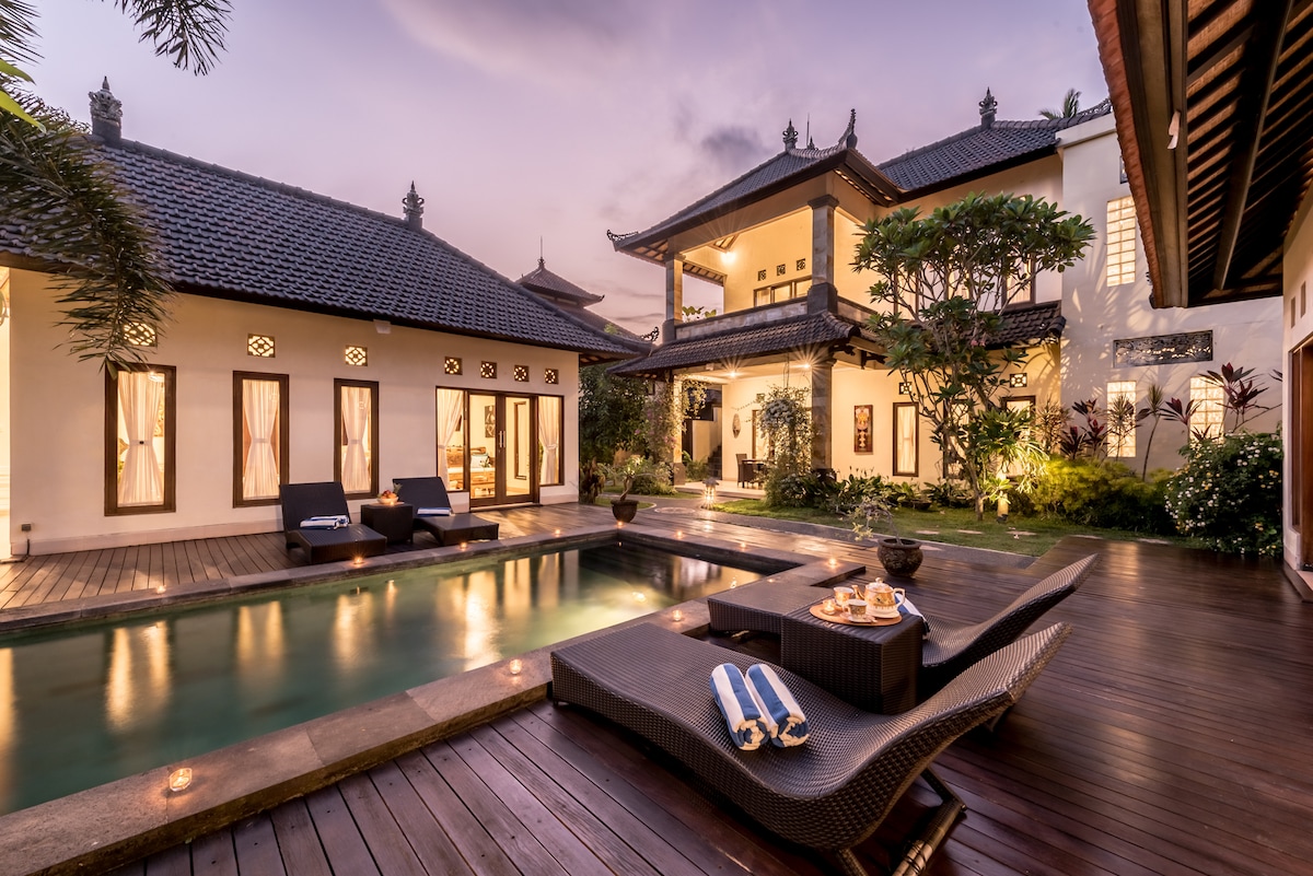 A charming exterior view of the villa showcases a serene swimming pool surrounded by wooden decking. Lounge chairs are placed nearby, with soft lighting highlighting the architectural details of the two-story structure and lush greenery in the evening ambiance.