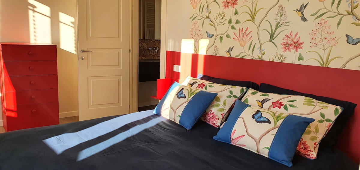 A bedroom is featured with a red accent wall adorned with floral wallpaper. A spacious bed is neatly dressed with dark bedding and decorative pillows displaying butterfly designs. Soft sunlight streams through a nearby window, casting gentle shadows across the room.